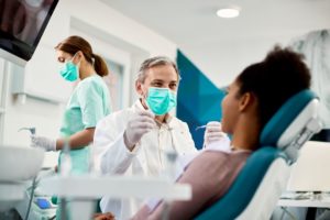 Picture in dental office with woman in the chair with a dentist in a green mask and white jacket approaching her while his assistant in green scrubs works behind him